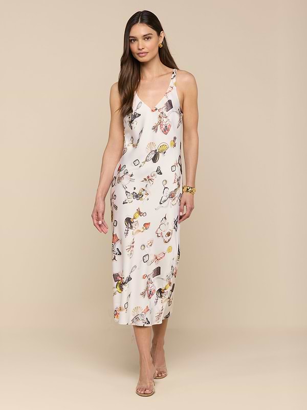 A woman stands against a plain white background wearing the Anika Slip Dress—a sleeveless, bias-cut satin white midi dress with a floral print—paired with strappy sandals.