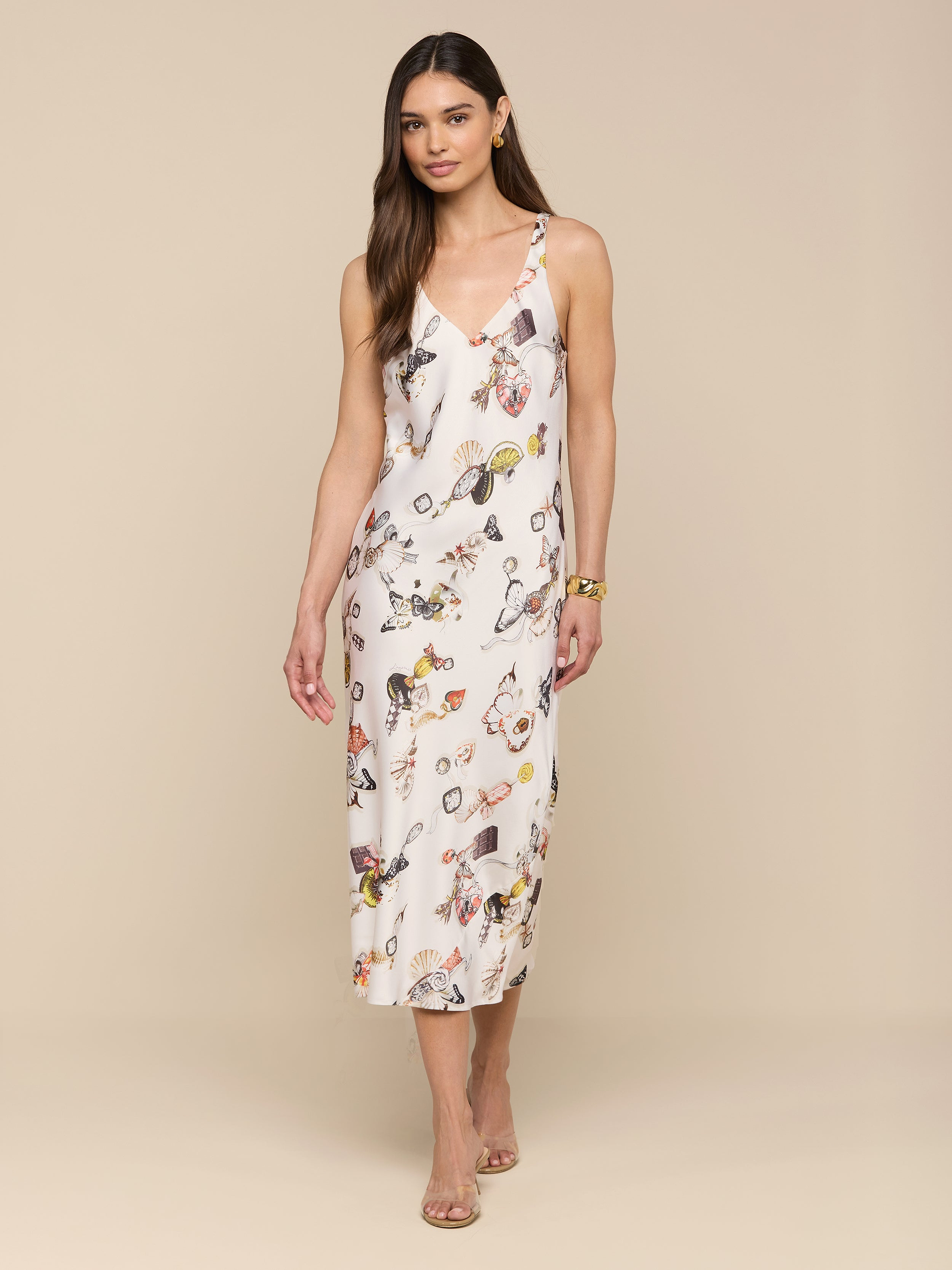 A woman stands against a plain white background wearing the Anika Slip Dress—a sleeveless, bias-cut satin white midi dress with a floral print—paired with strappy sandals.