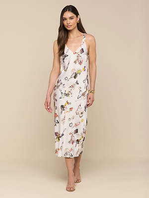 A woman with long brown hair models the Anika Slip Dress—a sleeveless, white satin midi slip dress with a colorful print. Styled with strappy heels and gold bracelets, she stands against a neutral background for an effortless evening look.