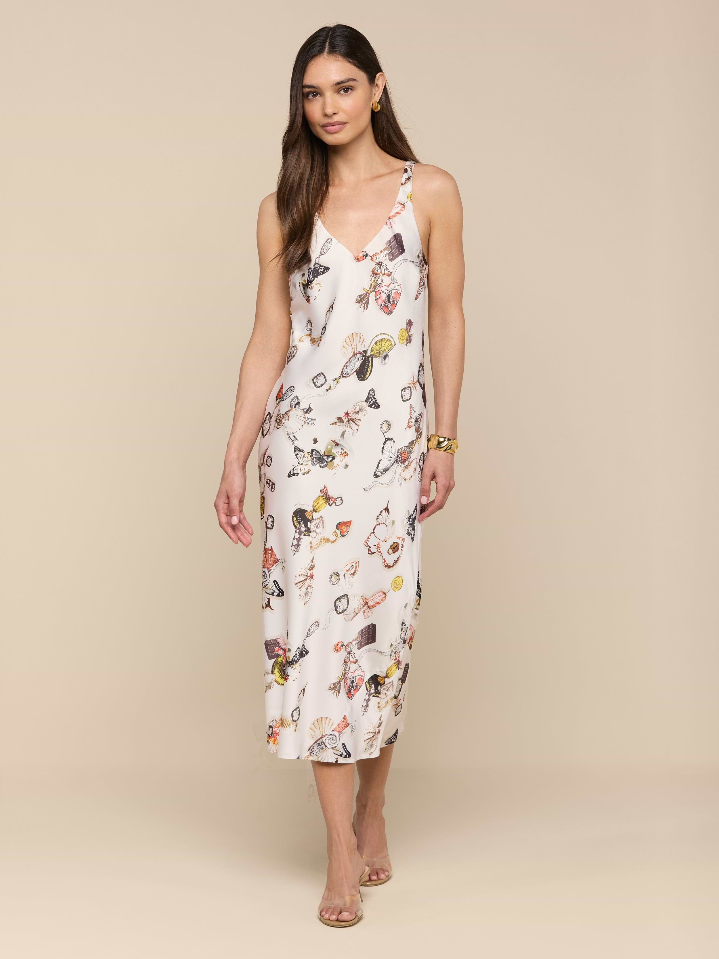 A woman stands against a plain white background wearing the Anika Slip Dress—a sleeveless, bias-cut satin white midi dress with a floral print—paired with strappy sandals.