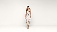 A woman with long brown hair wears a white, sleeveless, floral-patterned midi dress and nude sandals. She stands against a plain white background, facing forward with a relaxed expression.