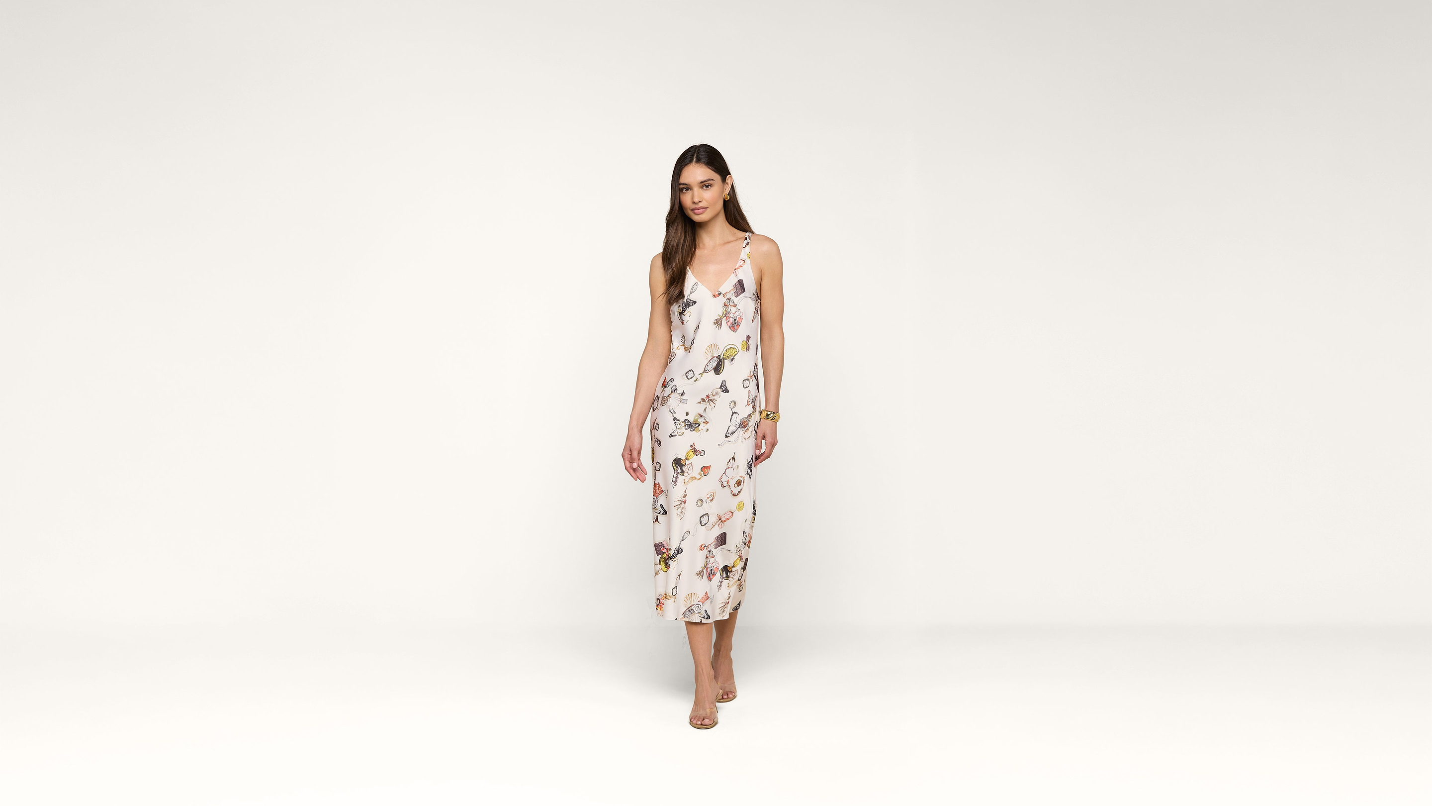 A woman with long brown hair wears a white, sleeveless, floral-patterned midi dress and nude sandals. She stands against a plain white background, facing forward with a relaxed expression.