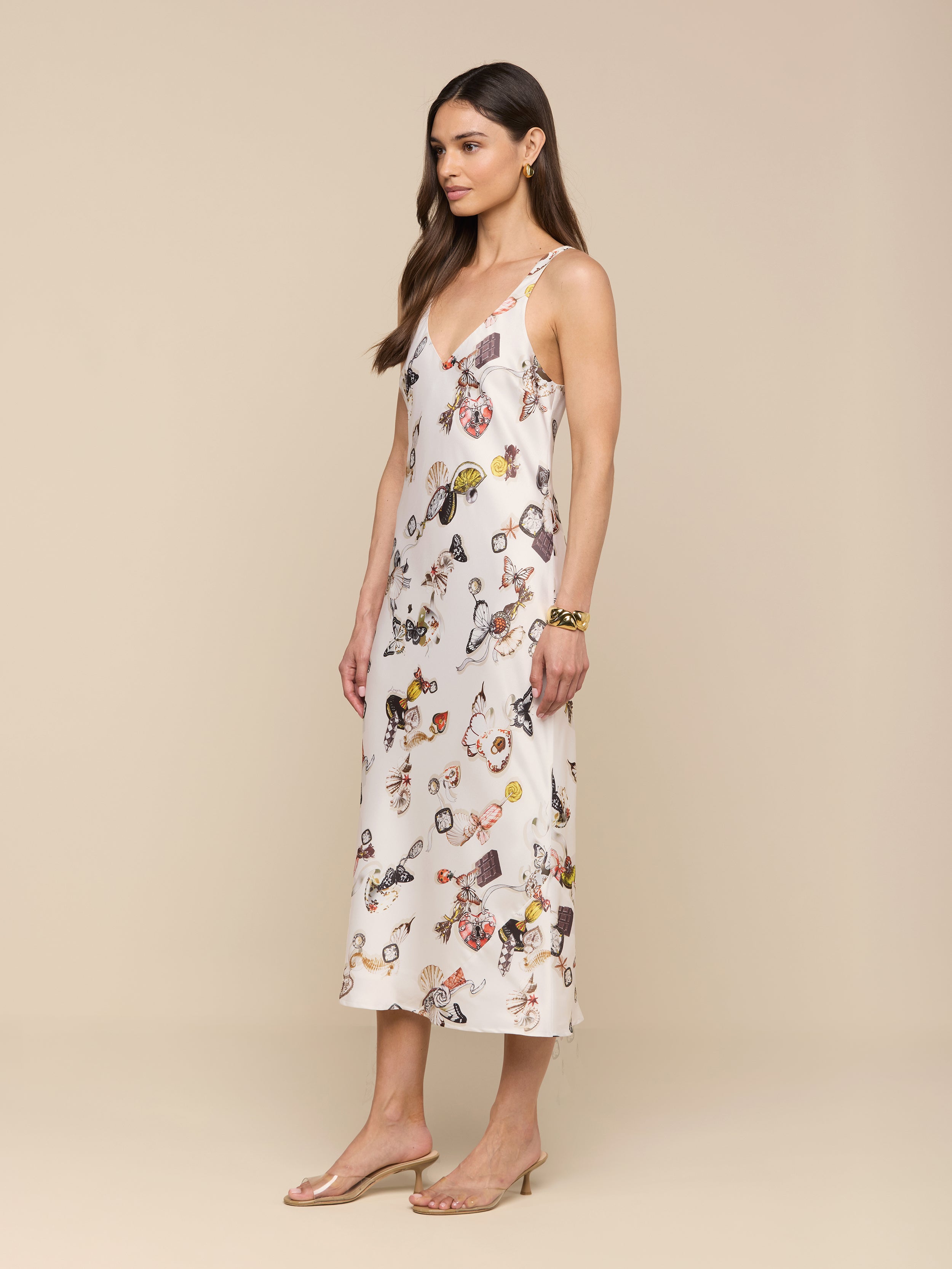 A woman stands in profile against a plain white background, wearing the Anika Slip Dress—a sleeveless, light-colored, ankle-length satin dress cut on the bias with a floral pattern—paired with sandals.