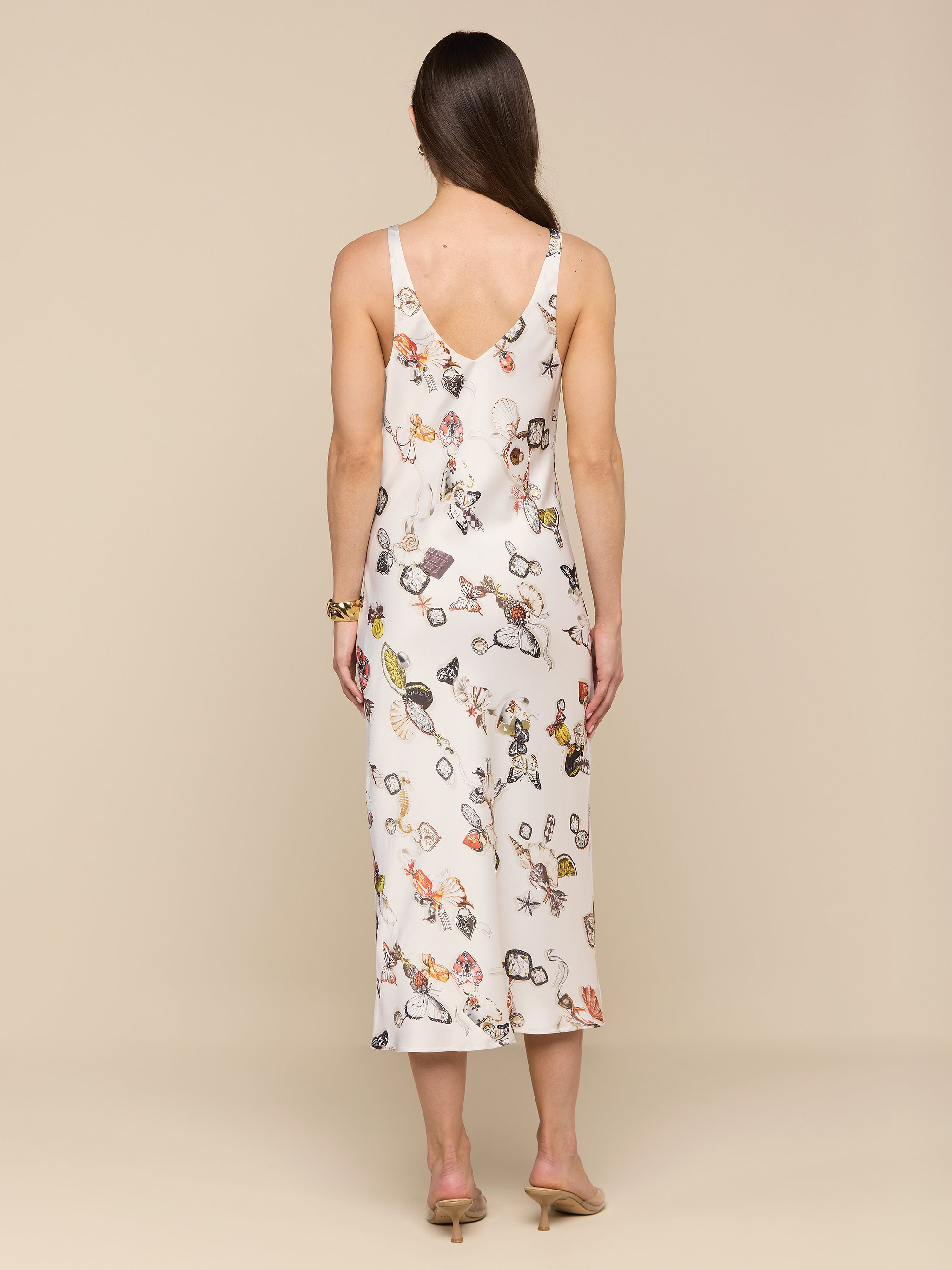 A woman with long brown hair stands facing away, wearing the Anika Slip Dress—a bias-cut satin slip dress featuring a floral pattern and a mid-length hem. She is also wearing heeled sandals, and the background is plain white.