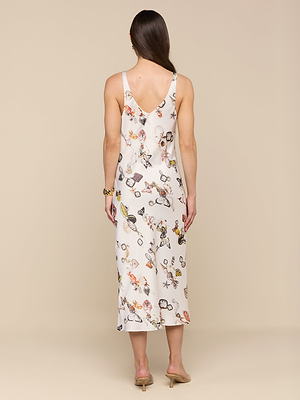 A woman with long brown hair faces away, wearing the Anika Slip Dress—a sleeveless, ankle-length white satin dress with colorful abstract patterns—paired with nude heeled sandals and a gold bracelet, against a beige background.