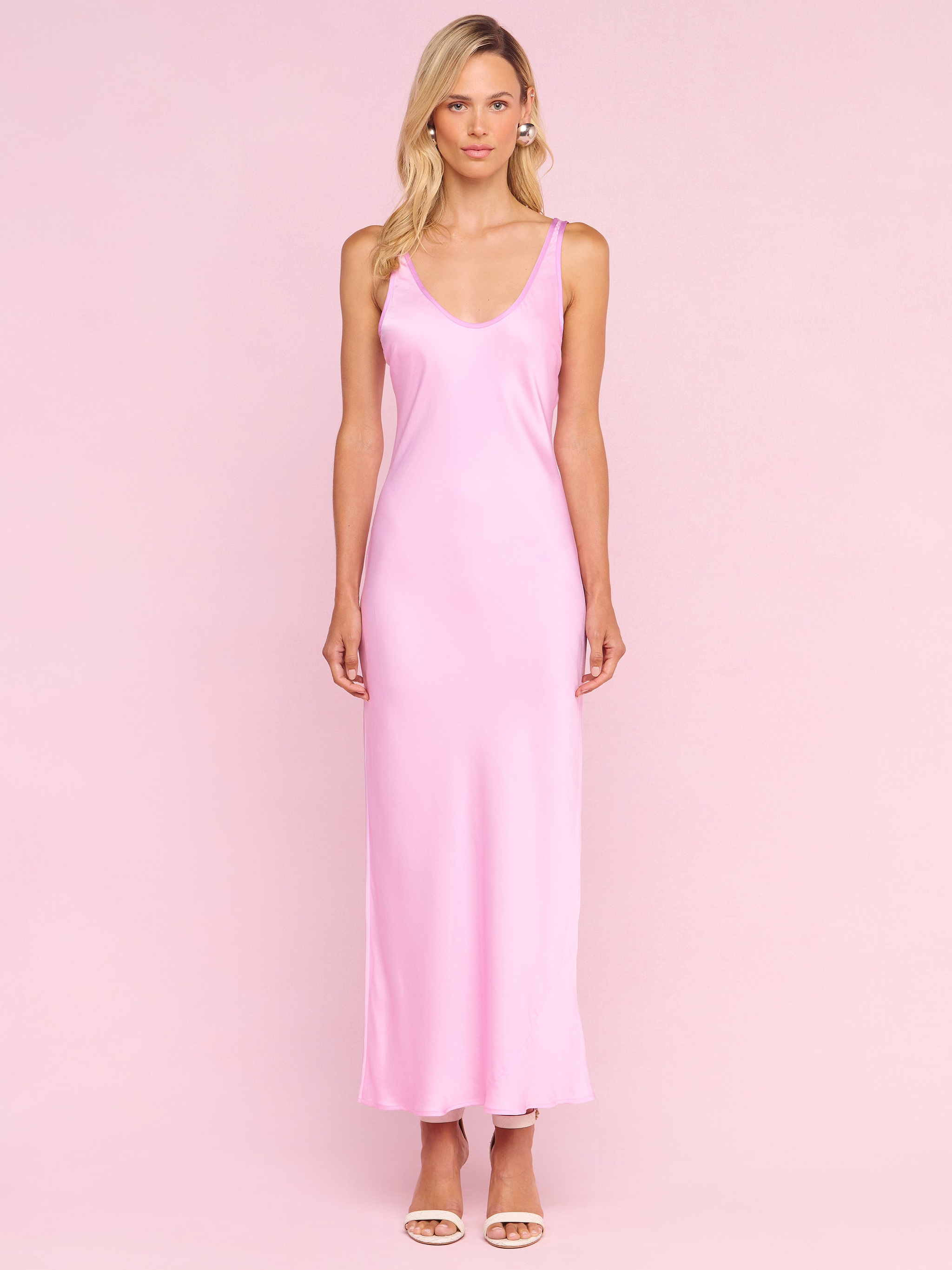 A woman with long blonde hair stands against a pale pink background wearing the Akiya Satin Maxi Dress—a sleeveless, floor-length pink satin dress—paired with open-toed nude heels and a neutral expression.