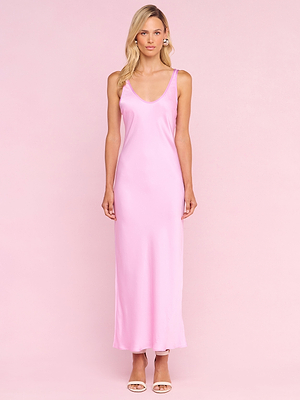 A woman with long blonde hair stands against a pale pink background wearing the Akiya Satin Maxi Dress—a sleeveless, floor-length pink satin dress—paired with open-toed nude heels and a neutral expression.