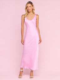 A woman with long blonde hair stands against a pale pink background wearing the sleeveless, ankle-length Akiya Satin Maxi Dress from the Legally Blonde collection and nude high-heeled sandals, looking slightly to the side.