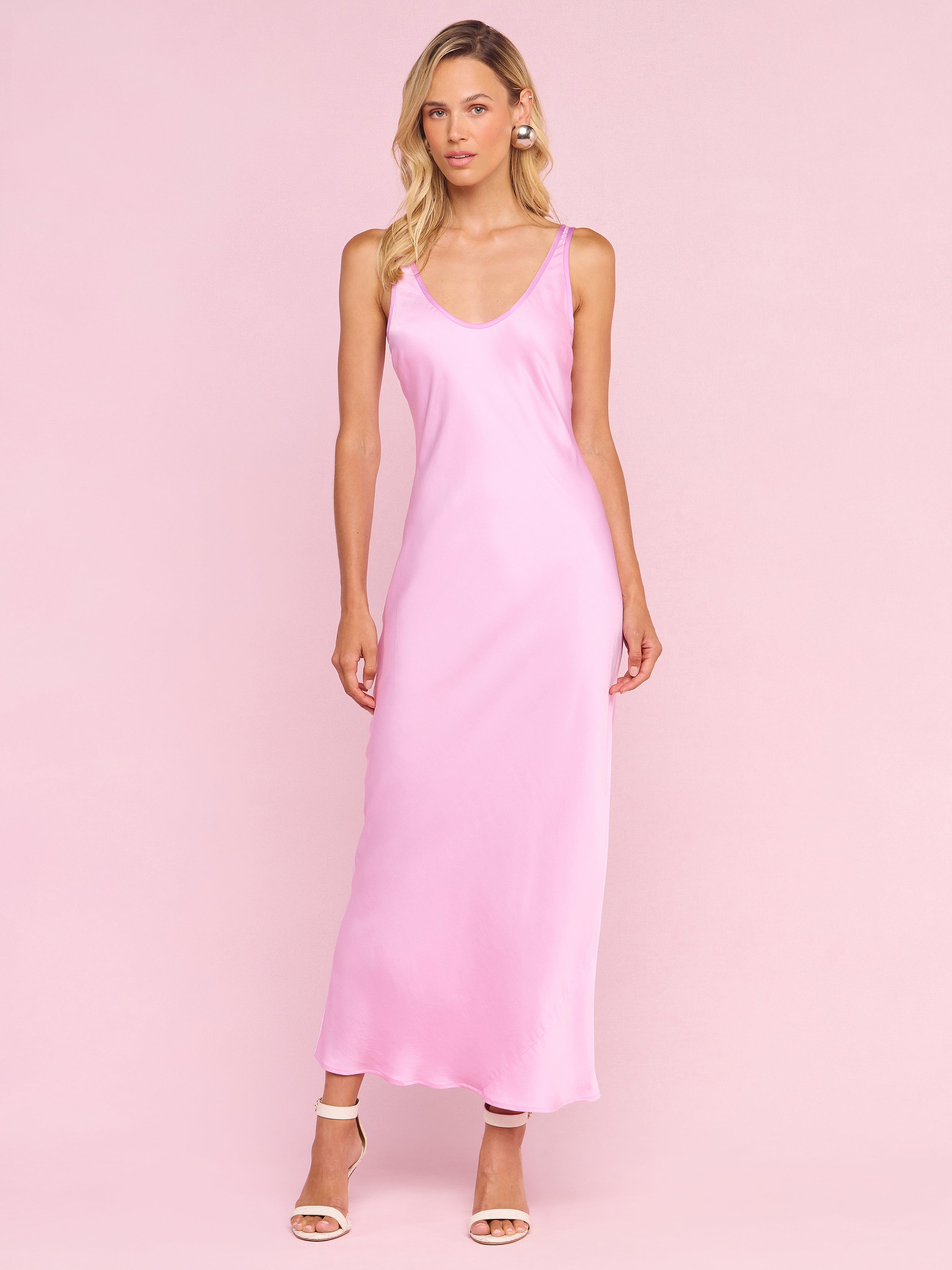 A woman with long blonde hair stands against a pale pink background wearing the sleeveless, ankle-length Akiya Satin Maxi Dress from the Legally Blonde collection and nude high-heeled sandals, looking slightly to the side.