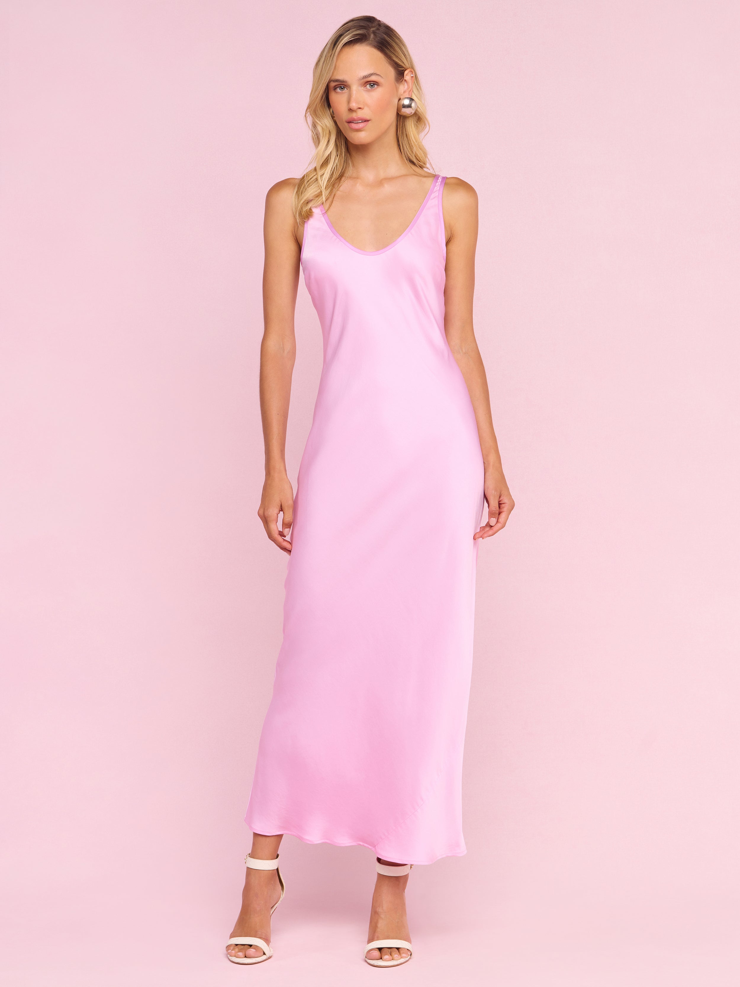 A woman with long blonde hair stands against a pale pink background wearing the sleeveless, ankle-length Akiya Satin Maxi Dress from the Legally Blonde collection and nude high-heeled sandals, looking slightly to the side.