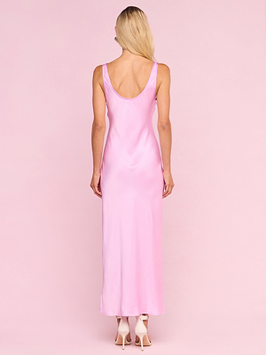 A woman with long blonde hair, seen from the back, wears a sleeveless Akiya Satin Maxi Dress and white heels against a pink background—a chic style from the Legally Blonde capsule collection.