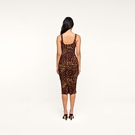 The woman with long dark hair wears the Anya Shirred Dress, a form-fitting leopard print midi dress, paired with black heels as she stands with her back to the camera against a plain white background.