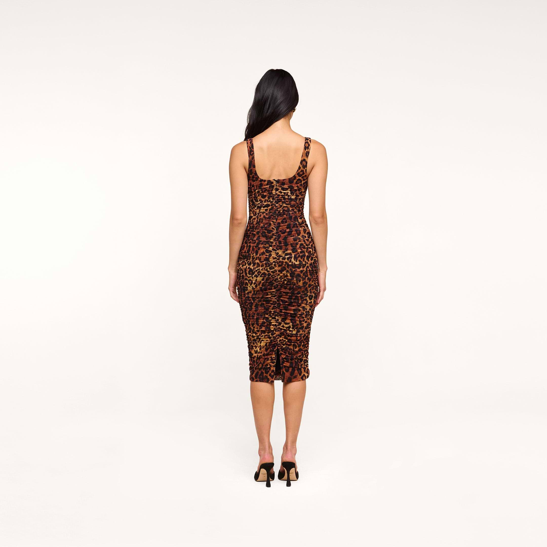 The woman with long dark hair wears the Anya Shirred Dress, a form-fitting leopard print midi dress, paired with black heels as she stands with her back to the camera against a plain white background.