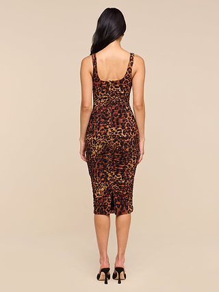 A woman stands with her back to the camera, wearing the Anya Shirred Dress—a sleeveless, form-hugging leopard print midi dress—paired with black heeled sandals, set against a plain beige background.