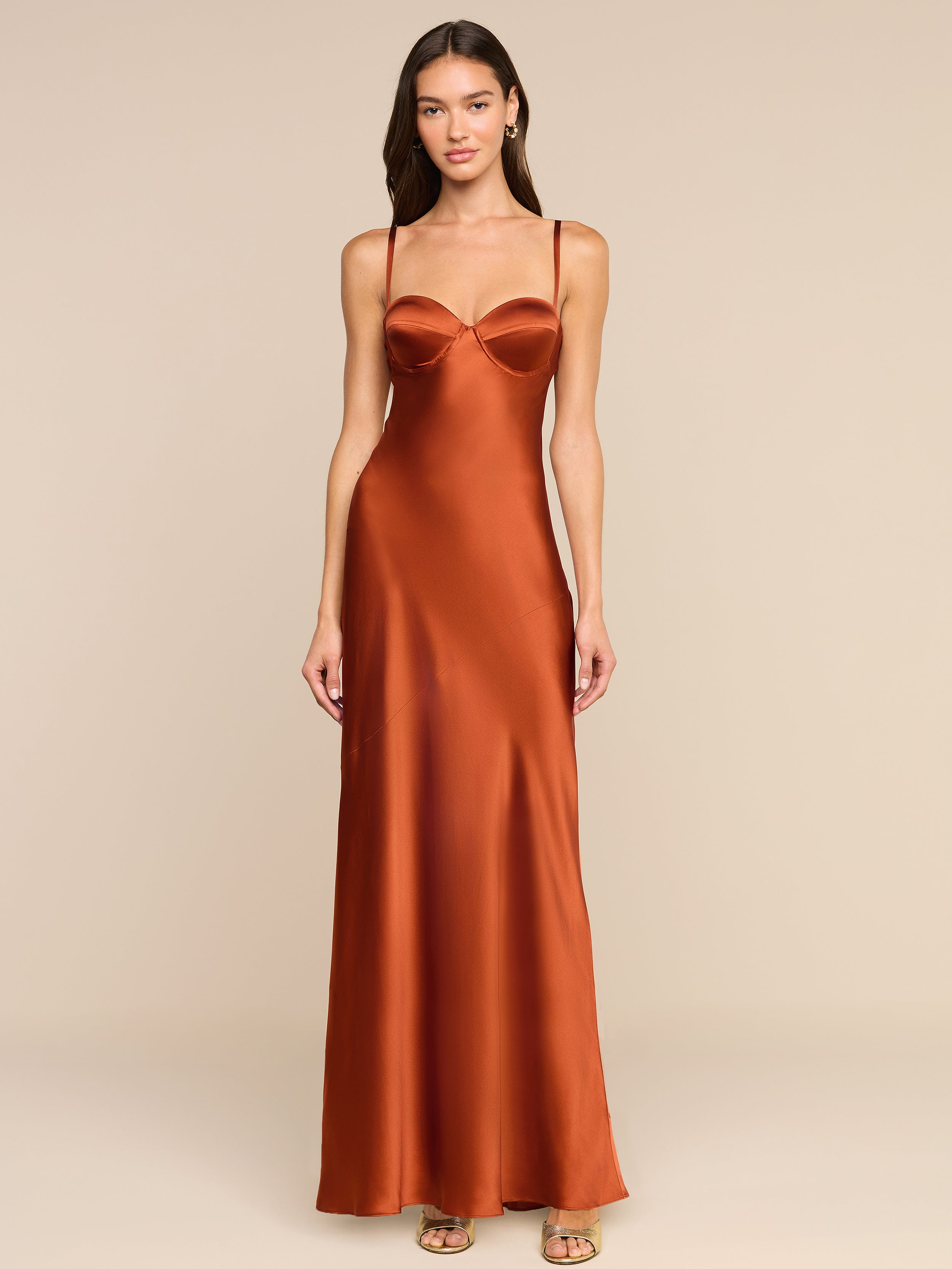 A woman stands facing forward against a plain white background, embodying vintage glamour in the Sayla Silk Gown—a long copper satin dress with delicate spaghetti straps.