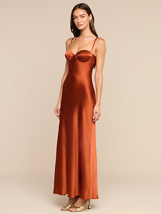Vêtue de la robe « Sayla Silk Gown », une robe nuisette soyeuse de couleur orange brûlé, longue jusquau sol et à fines bretelles, la femme lassocie à des sandales à talons hauts dorées, dégageant un charme vintage sur un fond beige uni.