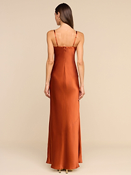 A woman stands facing away, wearing the Sayla Silk Gown—a long, copper silk dress with thin straps. The plain white background and her straight, loose hair cascading down her back create an event-ready look infused with vintage glamour.