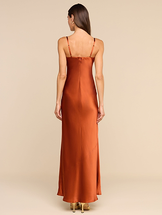 Une femme se tient dos à la caméra, rayonnant dun charme vintage dans la robe « Sayla Silk Gown » — une longue robe en satin orange brûlé à fines bretelles —, assortie à des talons hauts beiges, sur un fond beige uni.