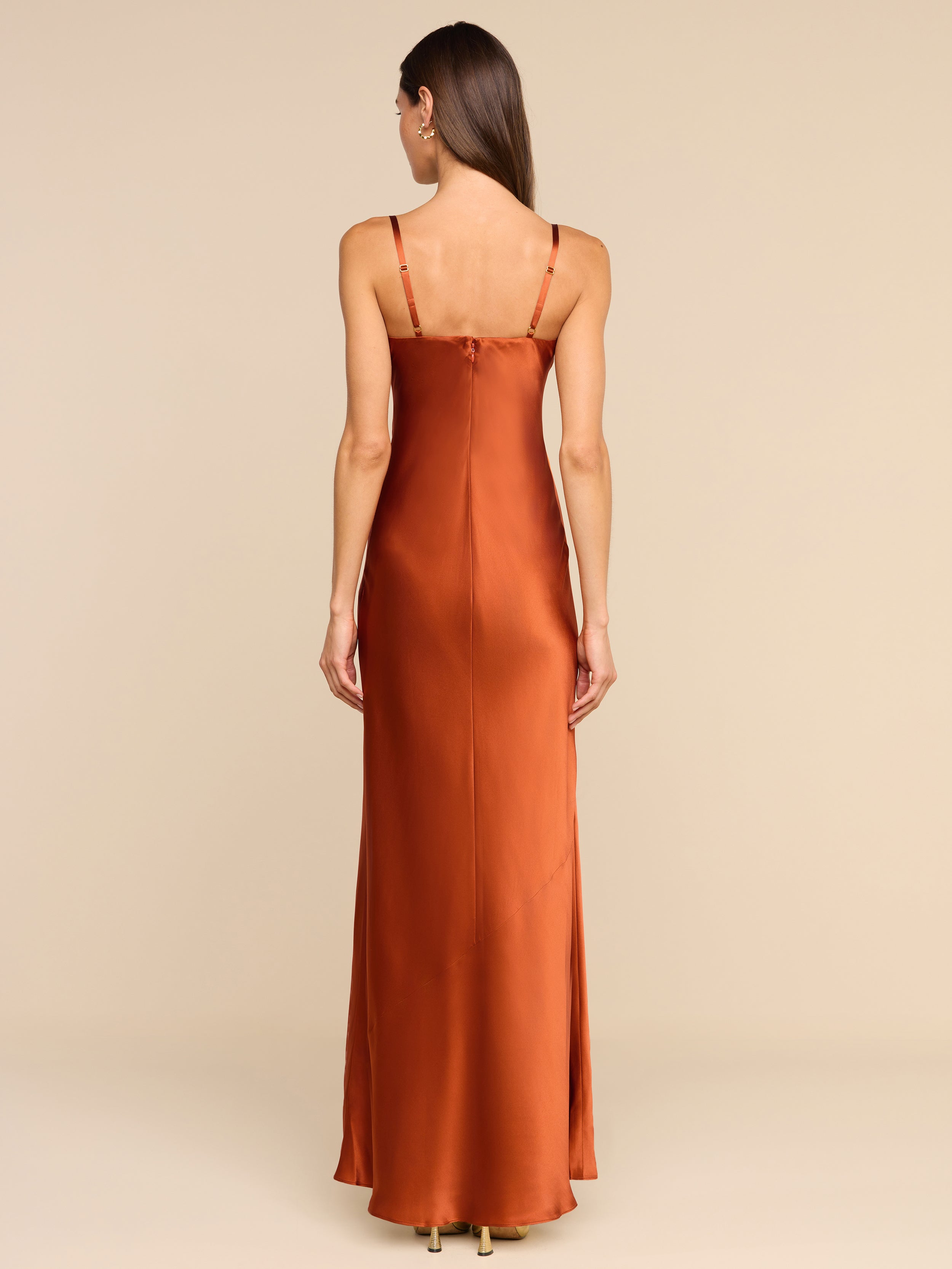 A woman stands facing away, wearing the Sayla Silk Gown—a long, copper silk dress with thin straps. The plain white background and her straight, loose hair cascading down her back create an event-ready look infused with vintage glamour.