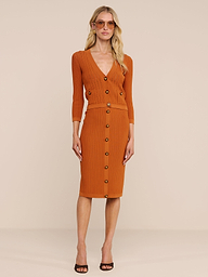 Eine Frau posiert vor einem schlichten Hintergrund in der Strickjacke „Irvin Pointelle“ und dem dazu passenden Rock. Der Look ist als leichte Übergangsmode mit schwarzen Pumps mit offener Spitze, Sonnenbrille, offenem Haar und einem neutralen Gesichtsausdruck gestylt.