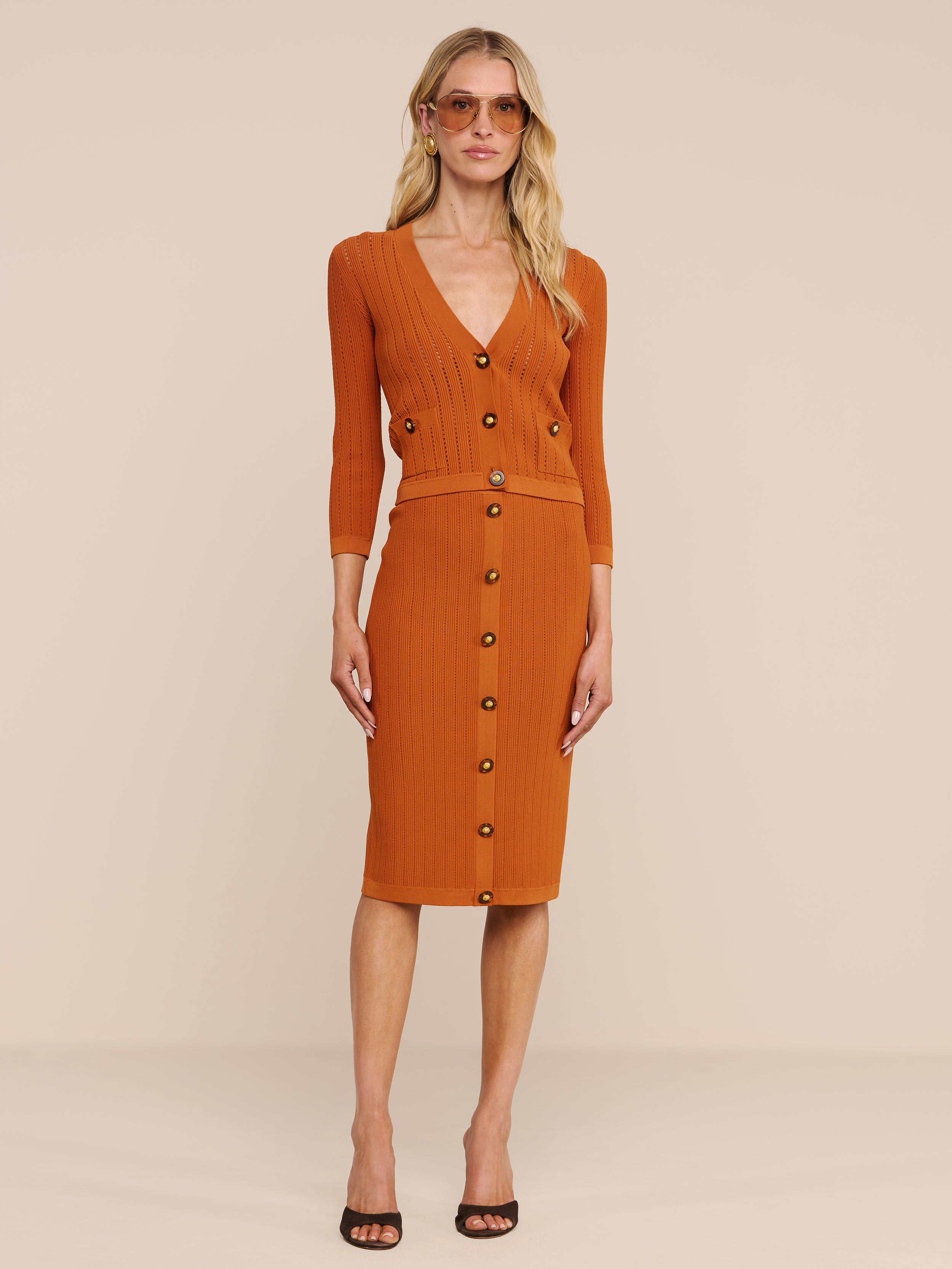Eine Frau posiert vor einem schlichten Hintergrund in der Strickjacke „Irvin Pointelle“ und dem dazu passenden Rock. Der Look ist als leichte Übergangsmode mit schwarzen Pumps mit offener Spitze, Sonnenbrille, offenem Haar und einem neutralen Gesichtsausdruck gestylt.