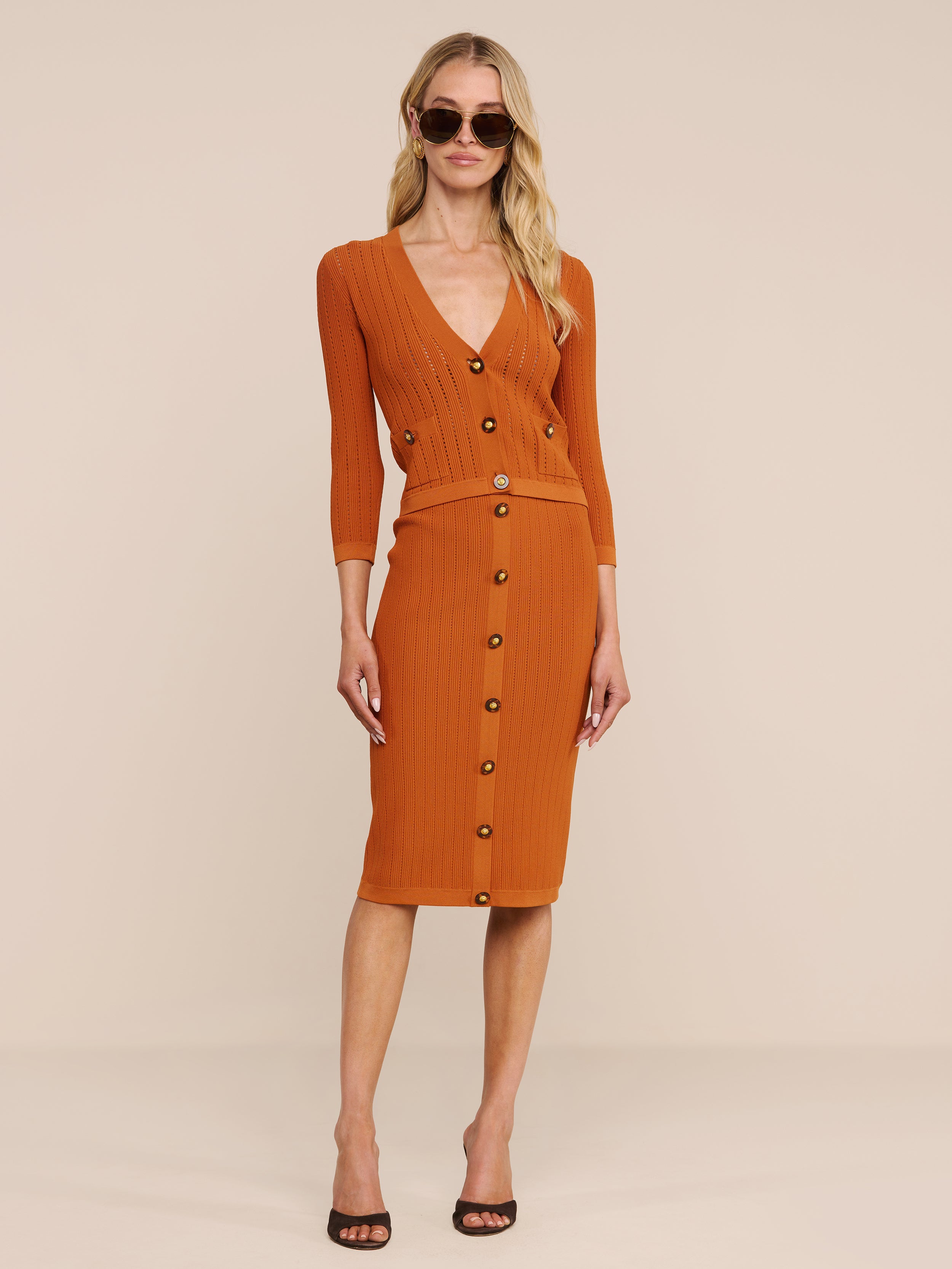 Eine Frau in einem orangefarbenen, gerippten Strickrock und der Strickjacke „Irvin Pointelle Knit Cardigan“ steht vor einem beigen Hintergrund. Mit einer Sonnenbrille, langen blonden Haaren und schwarzen Pumps mit offener Spitze rundet sie ihren schicken, saisonunabhängigen Lagenlook ab.