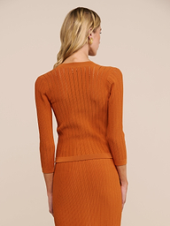 Eine Frau mit langen blonden Haaren trägt die Strickjacke „Irvin Pointelle“ über einem orangefarbenen, gerippten Oberteil mit Dreiviertelärmeln und einem dazu passenden Rock; die Aufnahme zeigt sie von hinten vor einem beigen Hintergrund.
