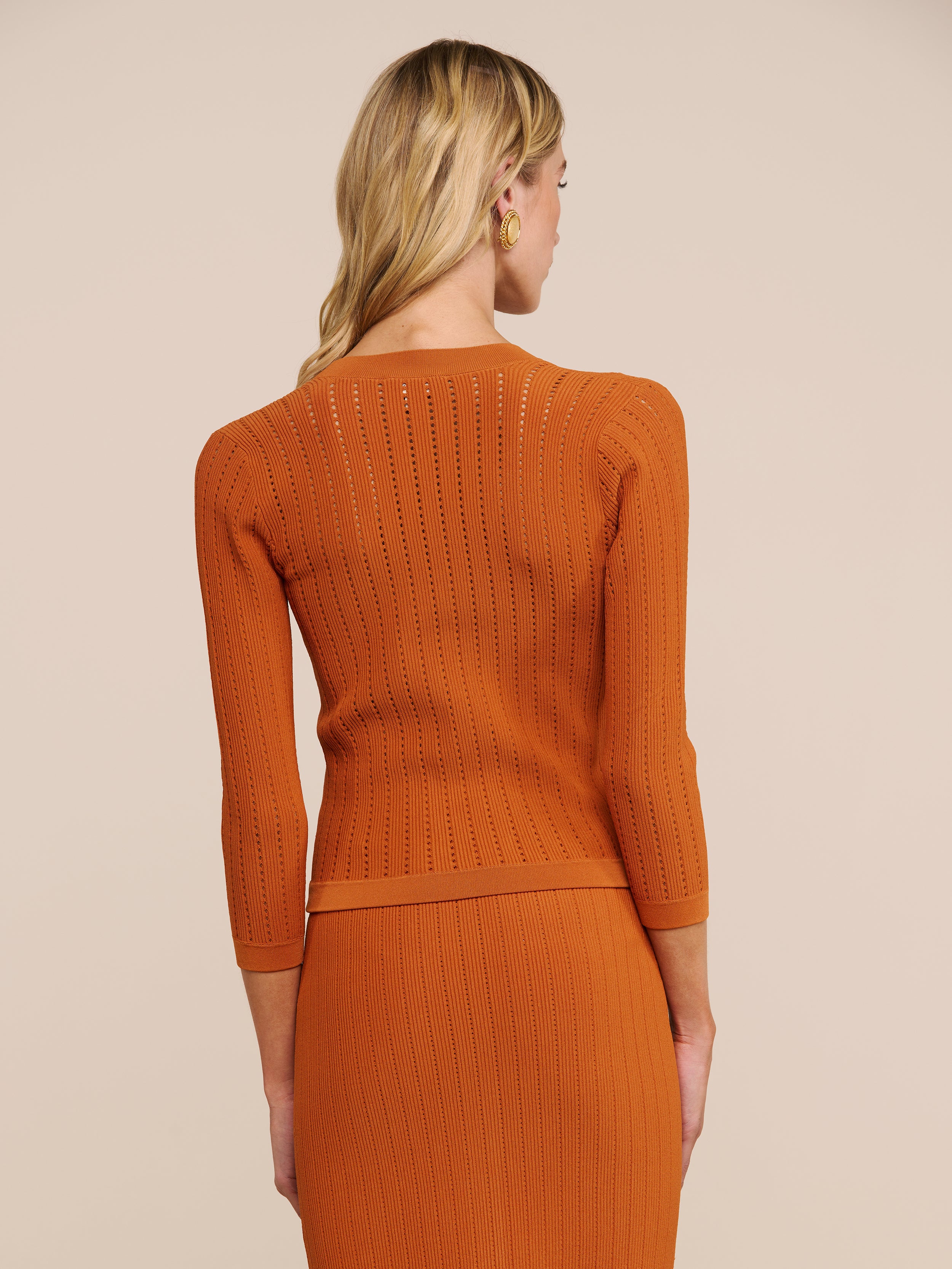 Eine Frau mit langen blonden Haaren trägt die Strickjacke „Irvin Pointelle“ über einem orangefarbenen, gerippten Oberteil mit Dreiviertelärmeln und einem dazu passenden Rock; die Aufnahme zeigt sie von hinten vor einem beigen Hintergrund.