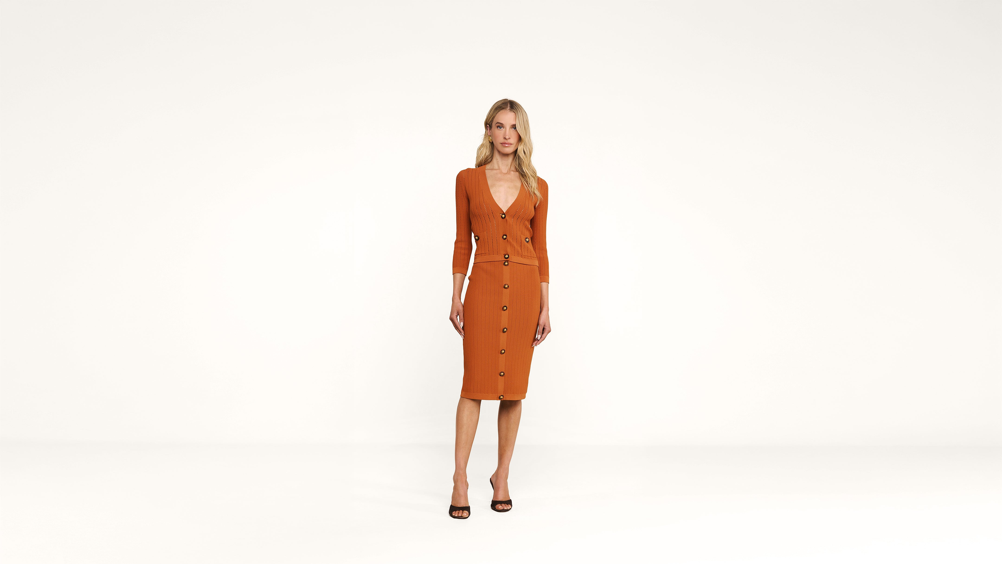 Woman stands in a minimal studio wearing an orange, button-down, fitted dress with long sleeves and black high-heeled sandals. Background is plain white.