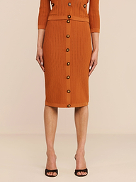 Ein Model präsentiert den Melania Pointelle-Strickrock in der Farbe „Rust“ mit gerippter Struktur, elastischem Bund und Knöpfen vorne, kombiniert mit schwarzen Pumps mit offener Spitze. Die figurbetonte Passform wird vor einem einfarbigen Hintergrund von den Schultern abwärts gezeigt.