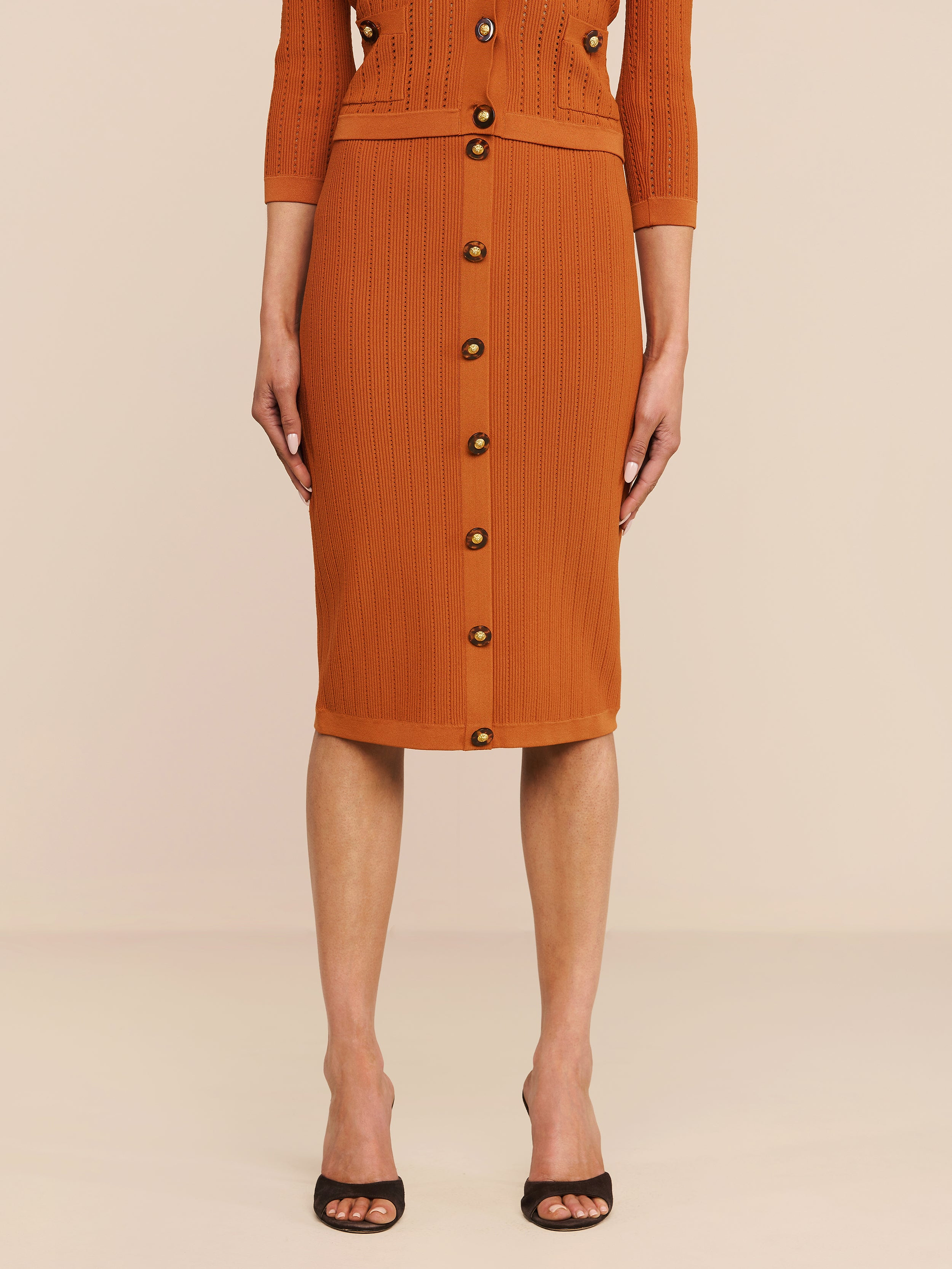 Ein Model präsentiert den Melania Pointelle-Strickrock in der Farbe „Rust“ mit gerippter Struktur, elastischem Bund und Knöpfen vorne, kombiniert mit schwarzen Pumps mit offener Spitze. Die figurbetonte Passform wird vor einem einfarbigen Hintergrund von den Schultern abwärts gezeigt.
