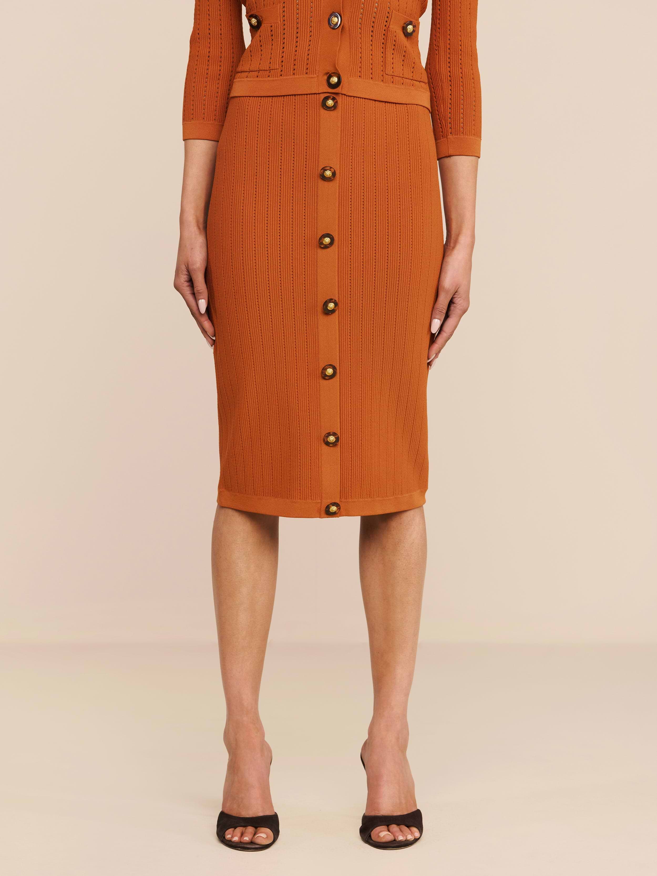 Ein Model präsentiert den Melania Pointelle-Strickrock in der Farbe „Rust“ mit gerippter Struktur, elastischem Bund und Knöpfen vorne, kombiniert mit schwarzen Pumps mit offener Spitze. Die figurbetonte Passform wird vor einem einfarbigen Hintergrund von den Schultern abwärts gezeigt.