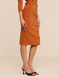Eine Frau steht im Profil und trägt den „Melania Pointelle Knit Skirt“ – einen rostfarbenen, figurbetonten Strickrock mit elastischem Bund –, kombiniert mit einem passenden Oberteil und schwarzen Sandalen mit offener Spitze und Absatz, vor einem neutralen Hintergrund.