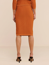Eine Person steht mit dem Rücken zur Kamera auf einem hellen Boden und trägt den orangefarbenen „Melania Pointelle Knit Skirt“ sowie ein passendes Oberteil und schwarze Pumps. Das Bild zeigt nur den unteren Rücken, die Beine und einen Teil der Arme der Person.