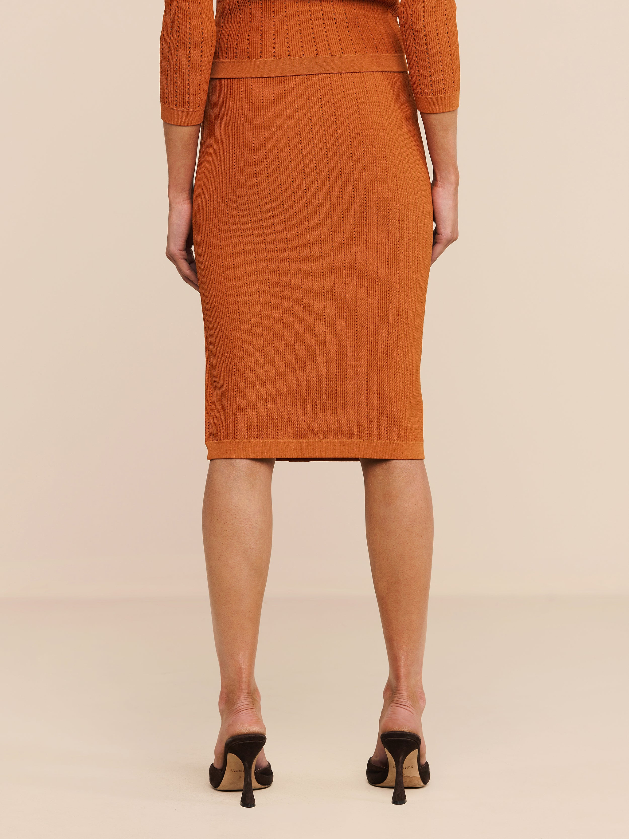 Eine Person steht mit dem Rücken zur Kamera auf einem hellen Boden und trägt den orangefarbenen „Melania Pointelle Knit Skirt“ sowie ein passendes Oberteil und schwarze Pumps. Das Bild zeigt nur den unteren Rücken, die Beine und einen Teil der Arme der Person.