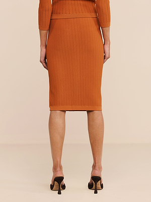 A person stands on a light floor, facing away, wearing the Melania Pointelle Knit Skirt in orange with a matching top and black heeled shoes. The image shows only their lower back, legs, and part of their arms.