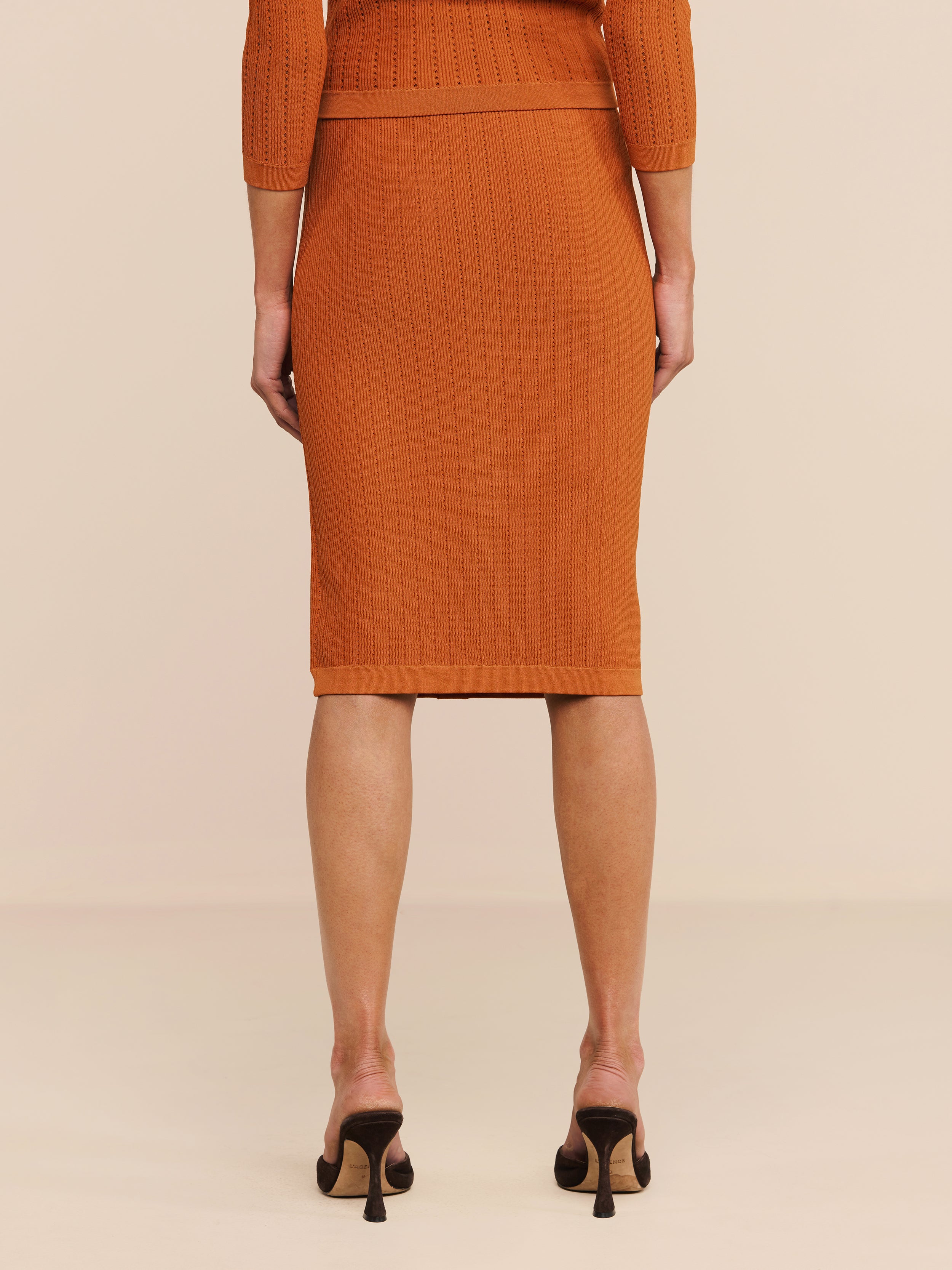 Eine Person steht mit dem Rücken zur Kamera auf einem hellen Boden und trägt den orangefarbenen „Melania Pointelle Knit Skirt“ sowie ein passendes Oberteil und schwarze Pumps. Das Bild zeigt nur den unteren Rücken, die Beine und einen Teil der Arme der Person.