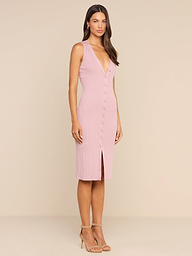 A woman stands in a studio wearing the Domino Dress—a sleeveless, light pink midi dress featuring a button-down front, V-neckline, and front slit. She has long hair and pairs the look with strappy high-heeled sandals.