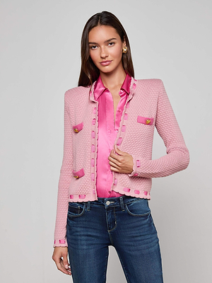 A woman with long brown hair models the L'AGENCE Astor Cardigan, a Parisian-inspired pink knit with decorative pockets, layered over a shiny pink blouse and blue jeans, standing against a plain light gray background.