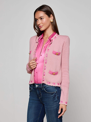 A woman in the L'AGENCE Astor Cardigan, a Parisian-inspired pink knit with gold buttons, layers it over a shiny pink blouse and blue jeans. She accessorizes with gold earrings and has long brown hair against a plain light backdrop.