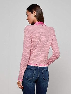 A woman with long brown hair stands facing away, wearing the L'AGENCE Astor Cardigan—a textured pink knit with decorative trim—paired with blue jeans, set against a plain light background for understated Parisian-inspired style.