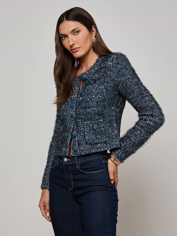 A woman with long brown hair wears the L'AGENCE Una Knit Cardigan—featuring multitone blue eyelash knit and patch pockets—with dark blue jeans, standing against a plain light gray background.