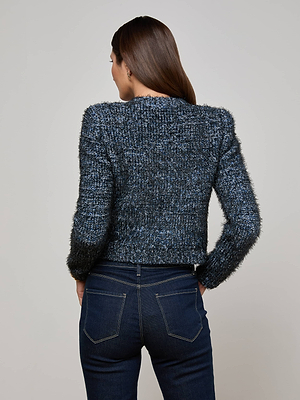 A woman with straight brown hair faces away, wearing the L'AGENCE Una Knit Cardigan in multitone blue and dark blue high-waisted jeans, standing against a plain light gray background.
