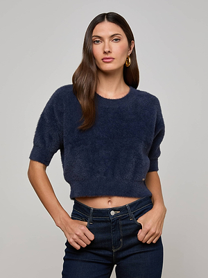 A woman with long brown hair models the L'AGENCE Guinevere Sweater, a plush knit navy blue cropped short-sleeve top, paired with dark jeans, standing hands on hips against a plain light gray background.