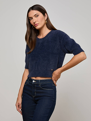 A woman with long brown hair models the L'AGENCE Guinevere Sweater, a navy plush knit short-sleeve top, paired with high-waisted dark blue jeans against a plain light gray background.