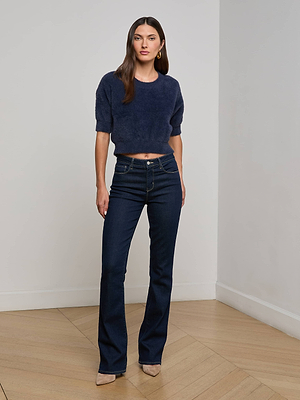 A woman with long brown hair poses indoors in the L’AGENCE Guinevere Sweater—navy, cropped, short-sleeve, plush knit—paired with dark blue high-waisted jeans and beige pointed-toe heels, set against a plain background.