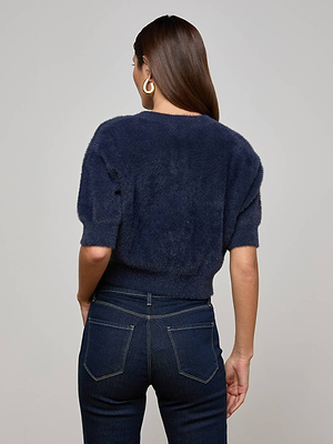 A woman with long brown hair and gold hoop earrings models the Guinevere Sweater by L'AGENCE—a plush knit navy blue cropped short-sleeve top—paired with dark blue jeans, facing away from the camera against a light gray background.