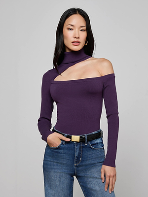 A woman with long dark hair models the L'AGENCE Azia Cutout Knit Top, an asymmetrical purple knit with one long sleeve and off-shoulder detail, styled with blue jeans and a black belt against a light gray background.