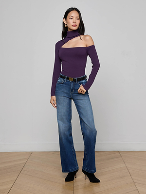 A woman with long dark hair and a neutral expression stands indoors against a plain wall, wearing the L'AGENCE Azia Cutout Knit Top in purple, blue flared jeans, a black belt, and black pointed-toe shoes.