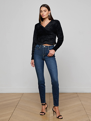 A woman stands indoors on a wooden floor, wearing the L'AGENCE Antonella Crossover Sweater with high-waisted blue jeans and black heels. She has straight brown hair, accessorized with earrings.